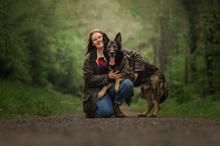 A woman crouches on a forest path with a German Shepherd dog beside her, wearing a dark olive jacket, blue jeans, and brown boots. The background is a blurred green woodland setting.
